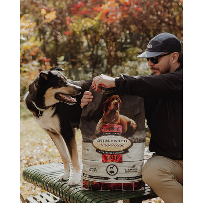 Man holding a bag of Oven-Baked Tradition dog food with a dog standing next to him outdoors.